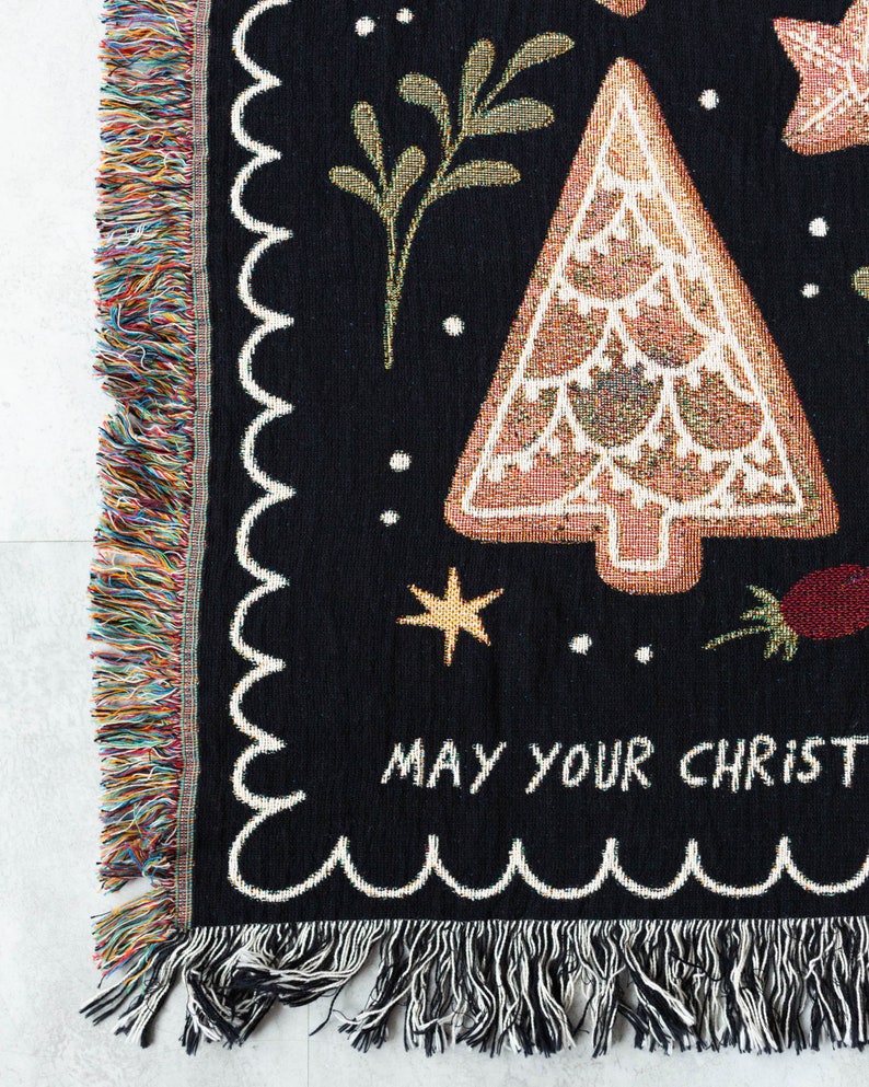 K&ouml;nnte beinhalten: Schwarze gewebte Wurfdecke mit einem Lebkuchenbaum-Design und dem Text "May your Christmas be merry and bright". Die Decke hat einen gewellten Rand und Fransen.