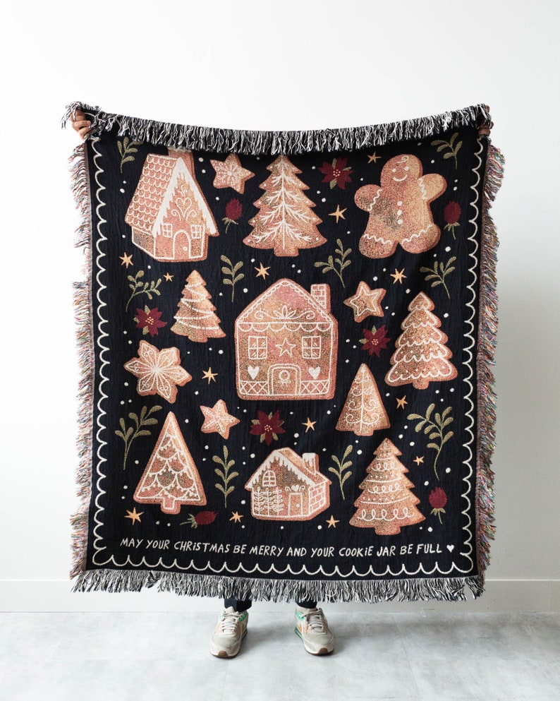 K&ouml;nnte beinhalten: Eine schwarze gewebte Wolldecke mit einem Lebkuchen-Motiv. Die Decke zeigt Lebkuchenh&auml;user, B&auml;ume und Sterne. Der Text "May your Christmas be merry and your cookie jar be full" ist unten auf der Decke aufgedruckt.
