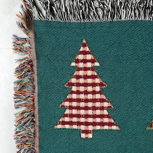 Op de afbeelding: Een groene geweven deken met een rood-wit geruit kerstboomontwerp. De deken heeft een franje rand met veelkleurige draden.
