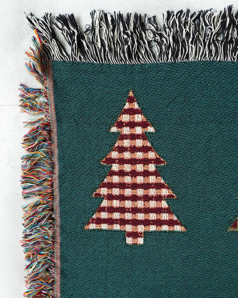 Op de afbeelding: Een groene geweven deken met een rood-wit geruit kerstboomontwerp. De deken heeft een franje rand met veelkleurige draden.