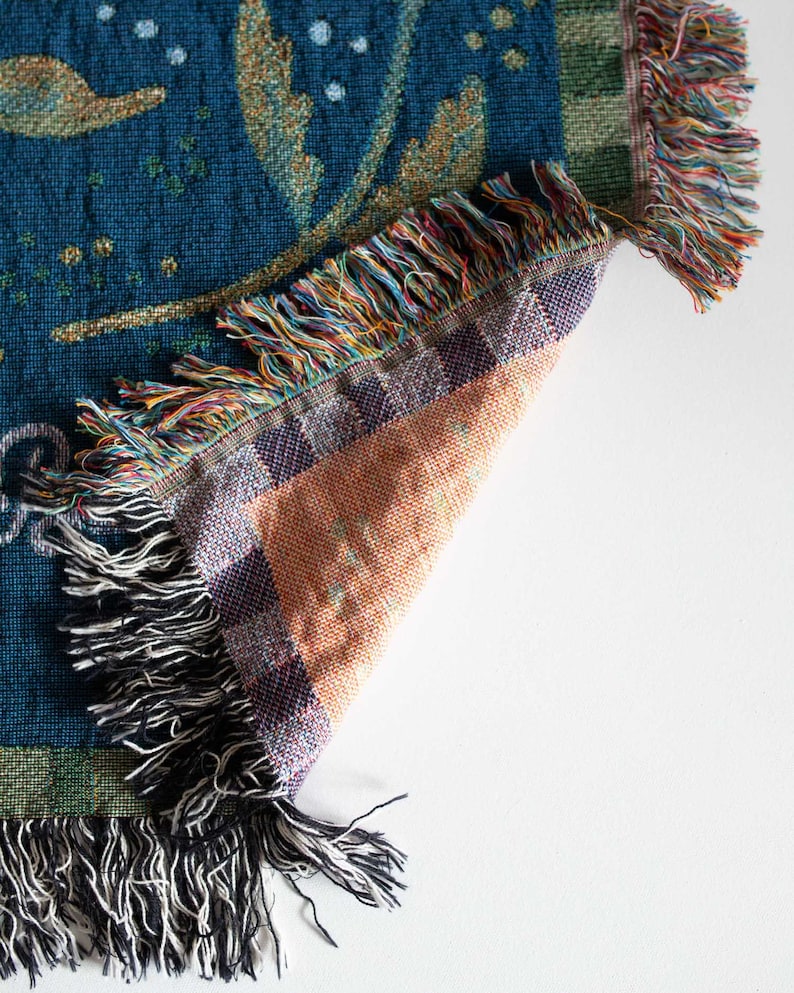 Pode incluir: Um cobertor tecido com fundo azul-petr&oacute;leo e design floral dourado. O cobertor tem uma borda com franjas com uma mistura de cores, incluindo azul-petr&oacute;leo, laranja e preto. O canto do cobertor est&aacute; dobrado, revelando a parte inferior.