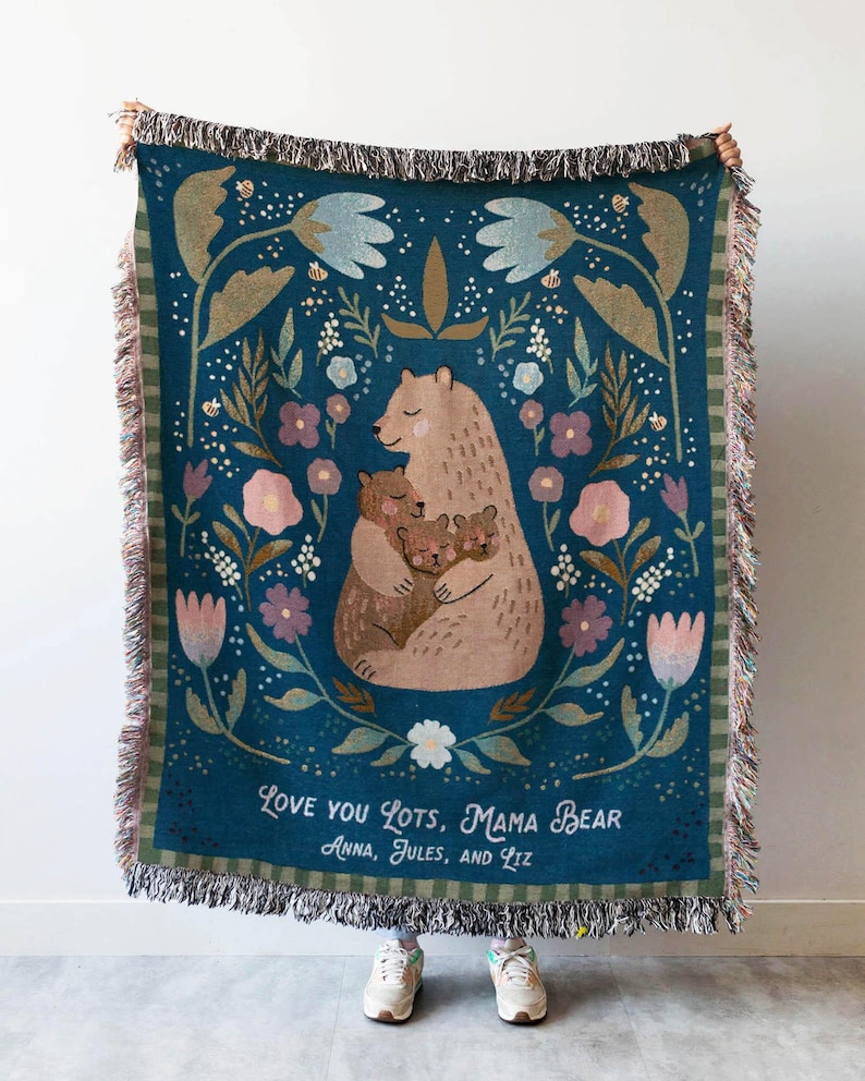 May include: A teal woven throw blanket with a bear family illustration and floral design. The blanket has a fringed edge and the text "Love You Lots, Mama Bear" with names below. The blanket is held up by a person.