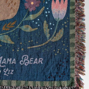 Pode incluir: Uma manta tecida com um fundo azul-petr&oacute;leo escuro, com desenhos florais e de urso. A manta tem uma borda com franjas multicoloridas. O texto na manta diz "Mama Bear" e "Liz".