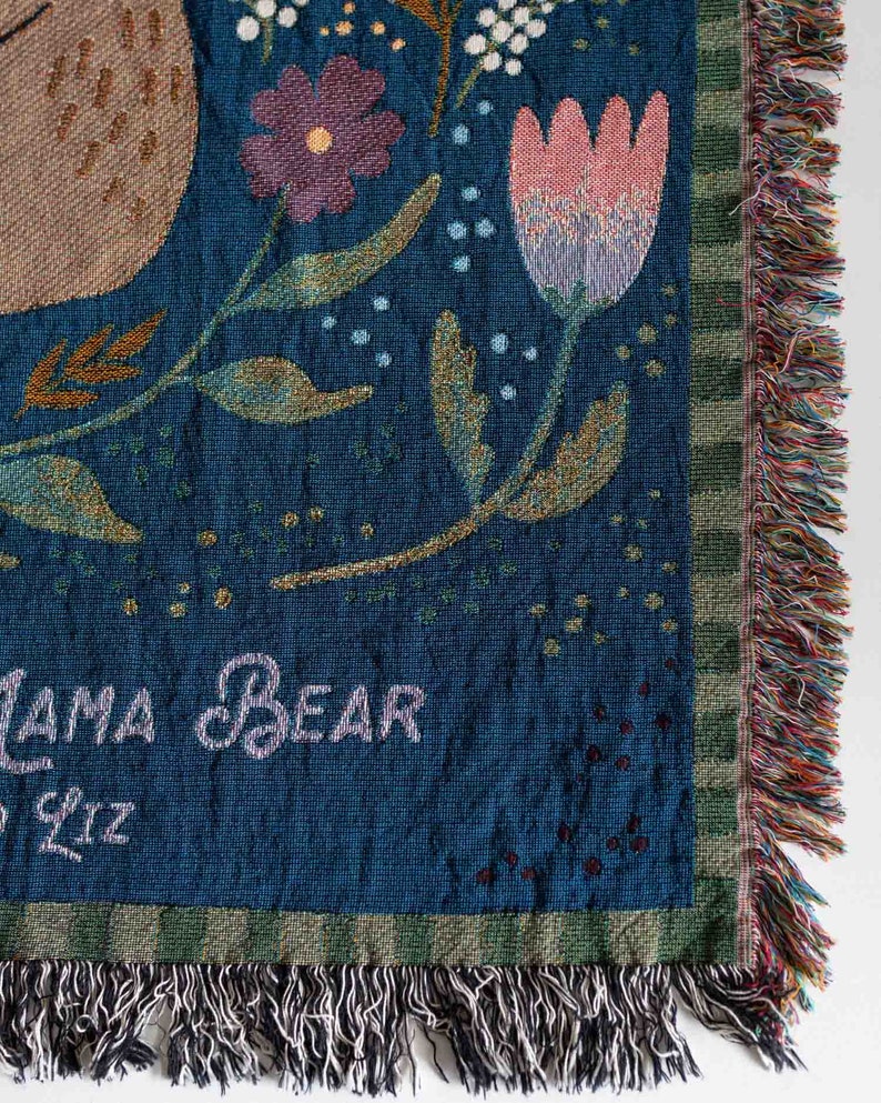 Pode incluir: Uma manta tecida com um fundo azul-petr&oacute;leo escuro, com desenhos florais e de urso. A manta tem uma borda com franjas multicoloridas. O texto na manta diz "Mama Bear" e "Liz".