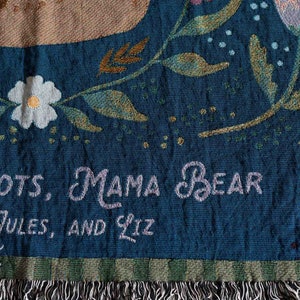 Pode incluir: Um cobertor tecido com fundo azul-petr&oacute;leo, com desenhos florais e de urso. O cobertor tem as palavras "MAMA BEAR" e nomes bordados. A borda inferior tem franjas.