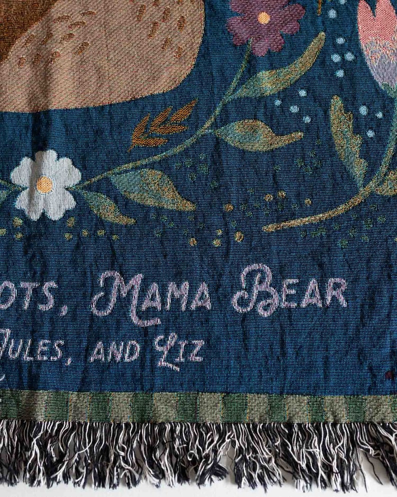 Pode incluir: Um cobertor tecido com fundo azul-petr&oacute;leo, com desenhos florais e de urso. O cobertor tem as palavras "MAMA BEAR" e nomes bordados. A borda inferior tem franjas.
