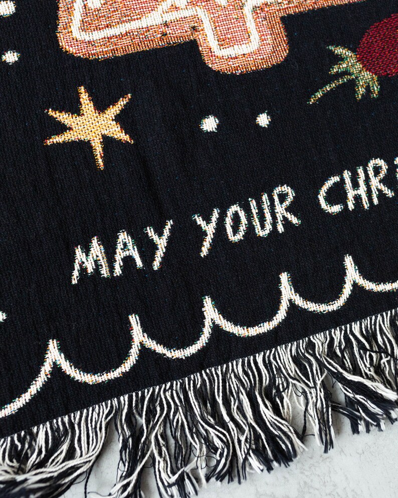 K&ouml;nnte beinhalten: Schwarze gewebte Wurfdecke mit wei&szlig;em Fransenrand. Die Decke zeigt ein farbenfrohes Design mit einem Stern, einer Palme und dem Text "MAY YOUR CHRISTMAS".