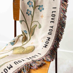 May include: A cream-colored woven throw blanket draped over a wooden chair. The blanket features a floral design with blue flowers and green leaves. The words "I LOVE YOU MOM" are printed in black along the border. The blanket has a fringed edge with multiple colors.