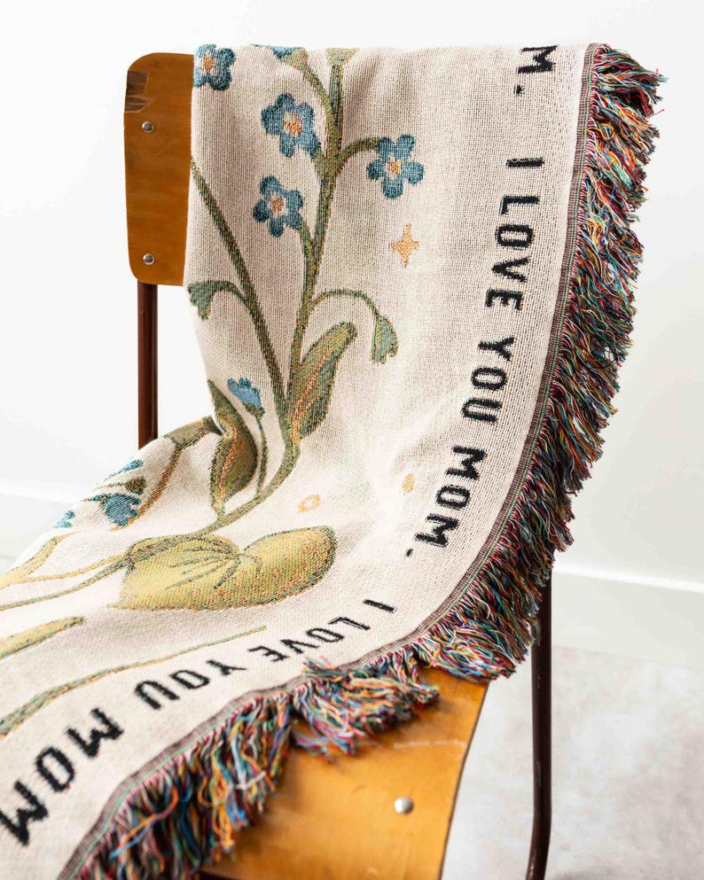May include: A cream-colored woven throw blanket draped over a wooden chair. The blanket features a floral design with blue flowers and green leaves. The words "I LOVE YOU MOM" are printed in black along the border. The blanket has a fringed edge with multiple colors.