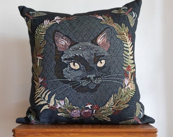 May include: A dark blue cushion featuring an embroidered black cat face in the centre. The cat's face is surrounded by a wreath of green leaves and flowers.