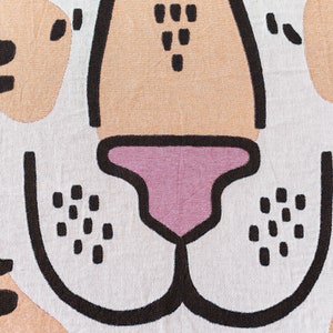May include: A woven throw blanket featuring a black and white illustration of a tiger's face. The tiger's nose is pink and the background is a light beige colour. The blanket has a black fringe.
