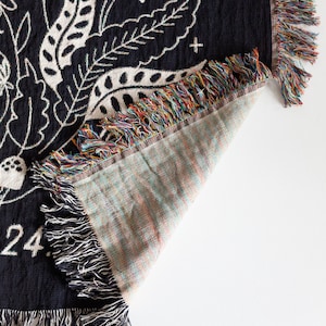 May include: Black woven throw blanket with a white floral design and fringe. The blanket is folded to show the back side, which is a light brown colour with a woven texture.