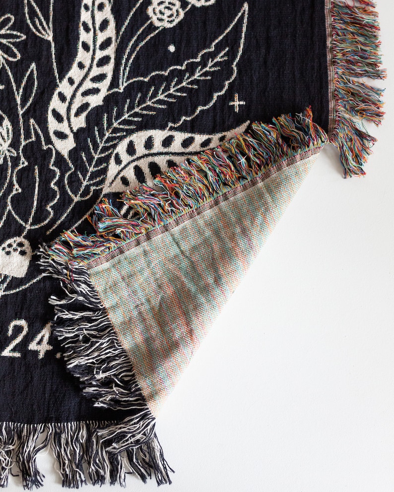 May include: Black woven throw blanket with a white floral design and fringe. The blanket is folded to show the back side, which is a light brown colour with a woven texture.
