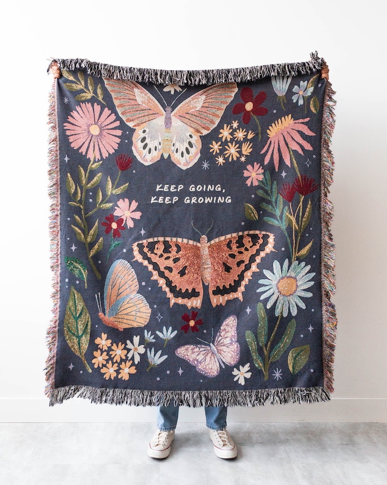 Woven blanket being held up by a person. Features a floral and butterfly design and the words "keep going, keep growing"