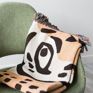 May include: A green armchair with a woven throw blanket featuring a geometric pattern in shades of brown, beige, and white. The blanket has fringed edges.
