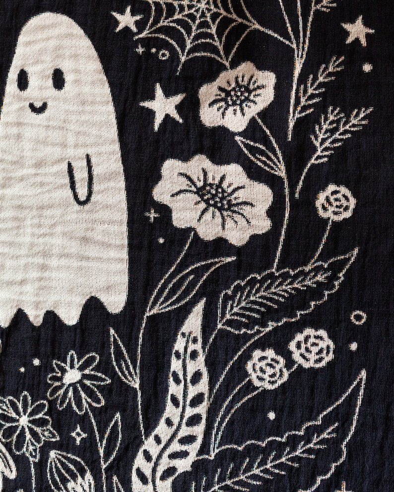 May include: Black fabric with a white embroidered design of a ghost, flowers, stars, and leaves. The ghost has a smiling face and a simple body shape.