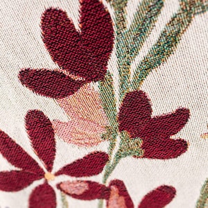 May include: Close-up of a woven fabric with a floral pattern. The design features deep red flowers with textured petals, set against a cream-colored background. Green and pink foliage accents the flowers, creating a detailed and colorful textile.