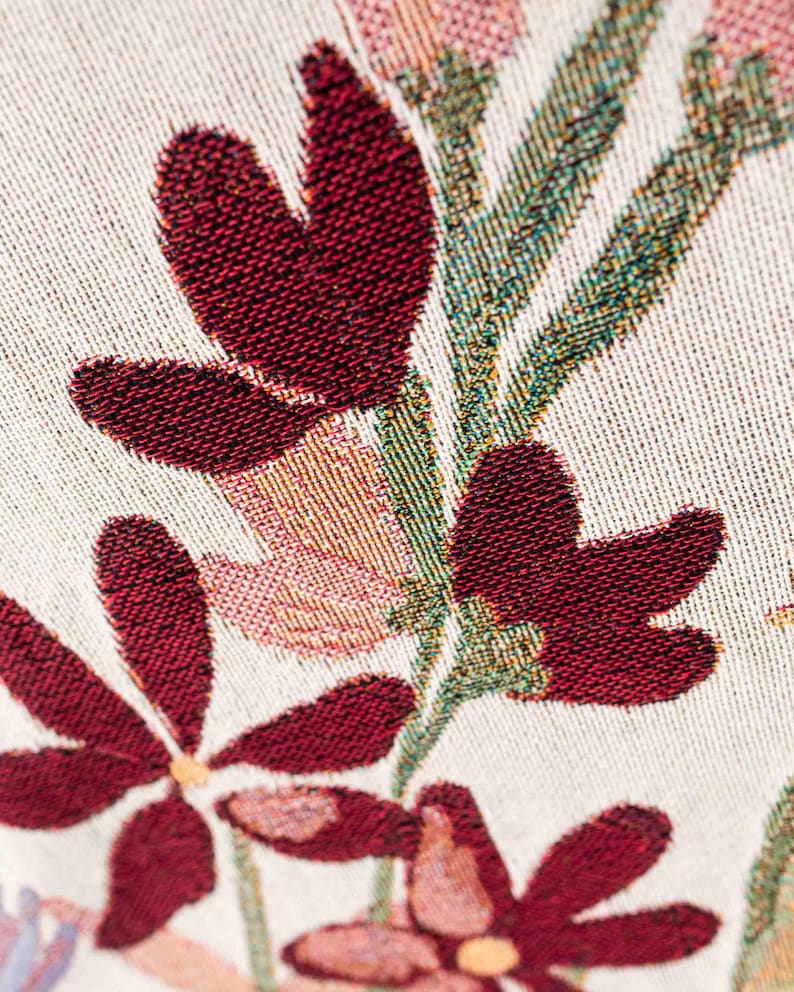 May include: Close-up of a woven fabric with a floral pattern. The design features deep red flowers with textured petals, set against a cream-colored background. Green and pink foliage accents the flowers, creating a detailed and colorful textile.