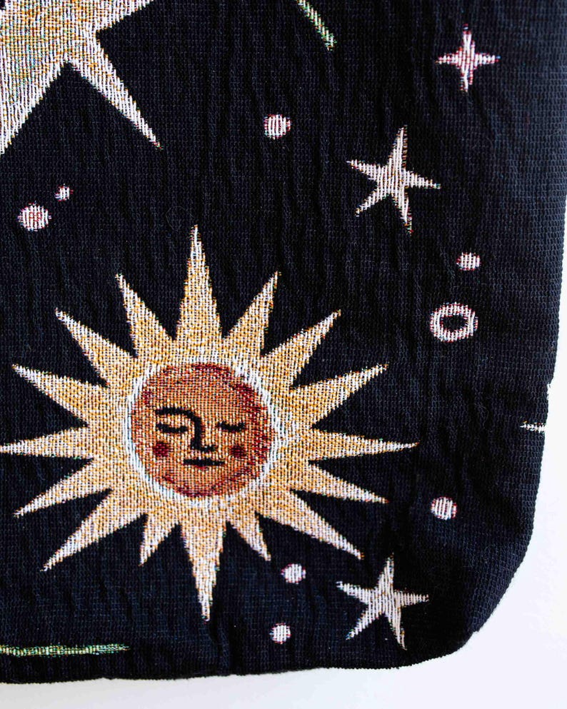 May include: Black fabric tote bag with a tapestry design of a sun with a face, stars, and small white dots.