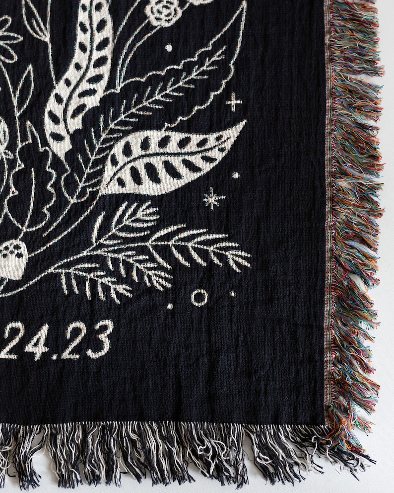 May include: Black woven throw blanket with a white floral design and the date "24.23" in the bottom left corner. The blanket has a fringe border.
