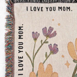 May include: Woven throw blanket with the text "I LOVE YOU MOM." Features floral designs in purple and orange, with green stems and leaves. The blanket has a fringed edge in multiple colors.