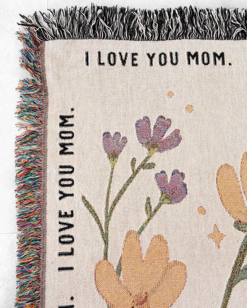May include: Woven throw blanket with the text "I LOVE YOU MOM." Features floral designs in purple and orange, with green stems and leaves. The blanket has a fringed edge in multiple colors.