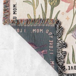 May include: Woven throw blanket with a floral design in shades of purple, pink, green, and cream. The blanket has a fringed edge with multiple colors. The text "I LOVE U MOM" is visible on the blanket.