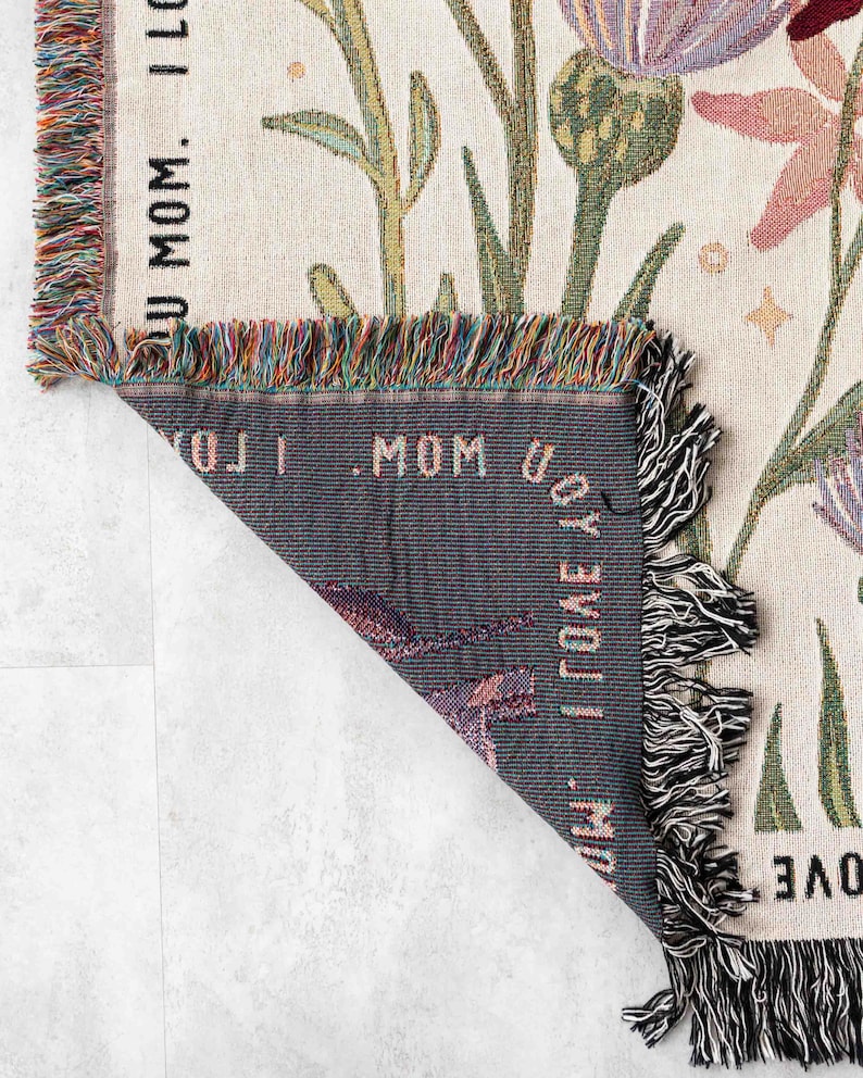 May include: Woven throw blanket with a floral design in shades of purple, pink, green, and cream. The blanket has a fringed edge with multiple colors. The text "I LOVE U MOM" is visible on the blanket.