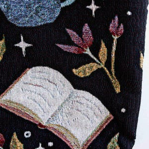 May include: A black tote bag with a tapestry design featuring a book, flowers, and stars. The book is open and has white pages. The flowers are red and yellow. The stars are white.