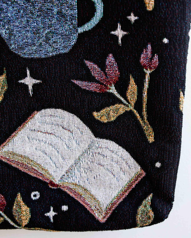 May include: A black tote bag with a tapestry design featuring a book, flowers, and stars. The book is open and has white pages. The flowers are red and yellow. The stars are white.