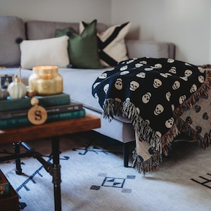 May include: A gray sectional sofa with a black and white skull patterned throw blanket draped over it. The sofa is decorated with pillows in shades of green and white. A wooden coffee table with a stack of books and a gold candle sits in front of the sofa.