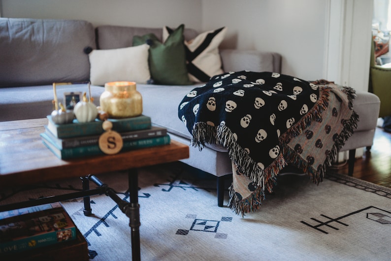 May include: A gray sectional sofa with a black and white skull patterned throw blanket draped over it. The sofa is decorated with pillows in shades of green and white. A wooden coffee table with a stack of books and a gold candle sits in front of the sofa.