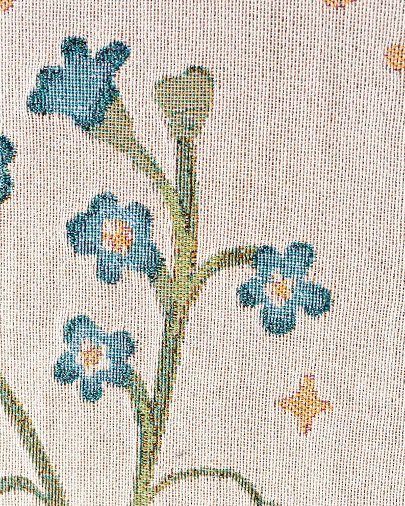 May include: Close-up of a woven fabric with a floral pattern. The design features blue flowers with yellow centers and green stems against a cream-colored background. Small gold star-shaped accents are also present.