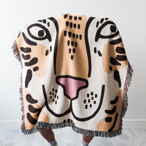 May include: A person is standing behind a woven tapestry throw blanket with a tiger face design. The blanket is beige, brown, and black with a pink nose. The person is wearing white trainers.