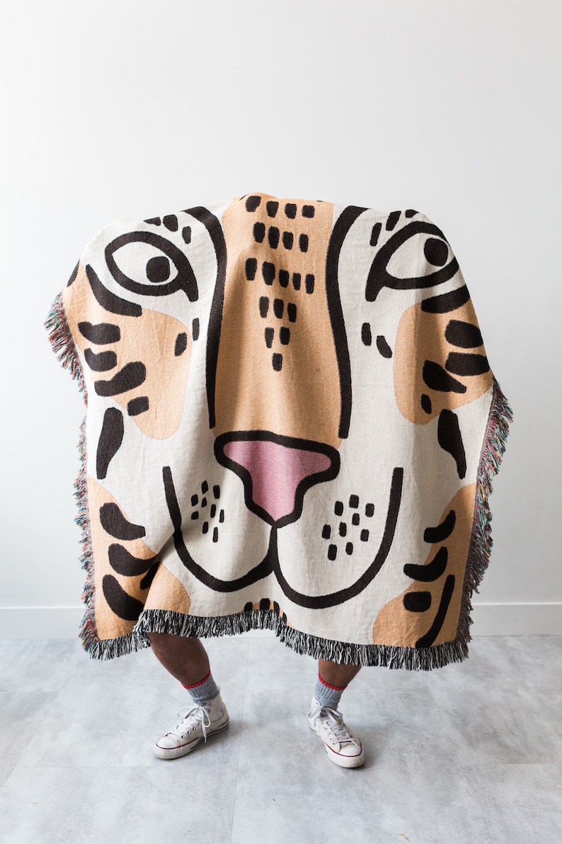 May include: A person is standing behind a woven tapestry throw blanket with a tiger face design. The blanket is beige, brown, and black with a pink nose. The person is wearing white trainers.