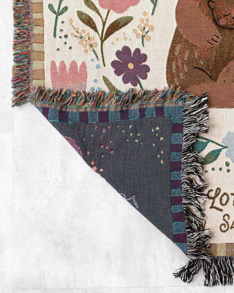 May include: A woven throw blanket featuring a floral design and a brown bear. The blanket has a fringed edge and the text "Lots of love" is visible in the bottom right corner.