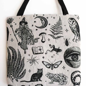 May include: A beige tote bag with black fabric handles featuring a repeating black ink illustration pattern of a witch with a broom, a crescent moon, ferns, an owl, a book, a frog, a spider, a moth, a cat, a toad, a snake, and an eye.