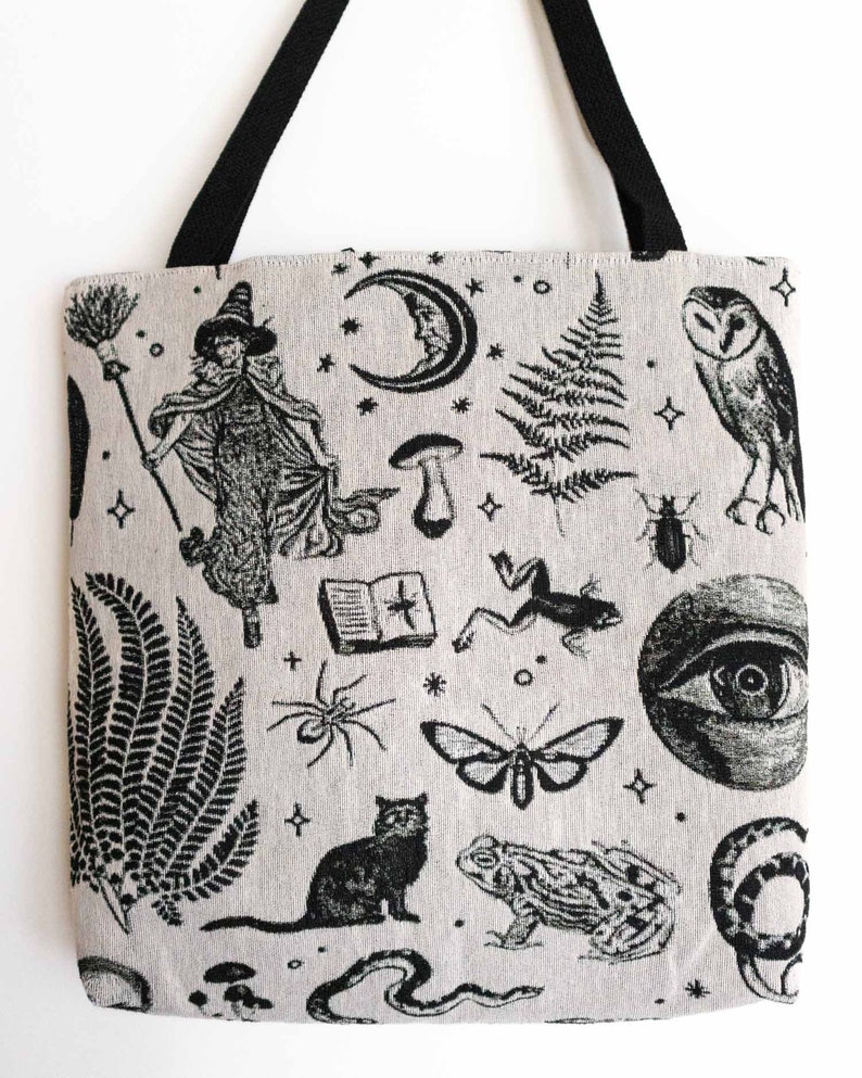 May include: A beige tote bag with black fabric handles featuring a repeating black ink illustration pattern of a witch with a broom, a crescent moon, ferns, an owl, a book, a frog, a spider, a moth, a cat, a toad, a snake, and an eye.