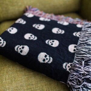 May include: A black throw blanket with a white skull pattern and fringe edges. The blanket is folded and placed on a green couch.