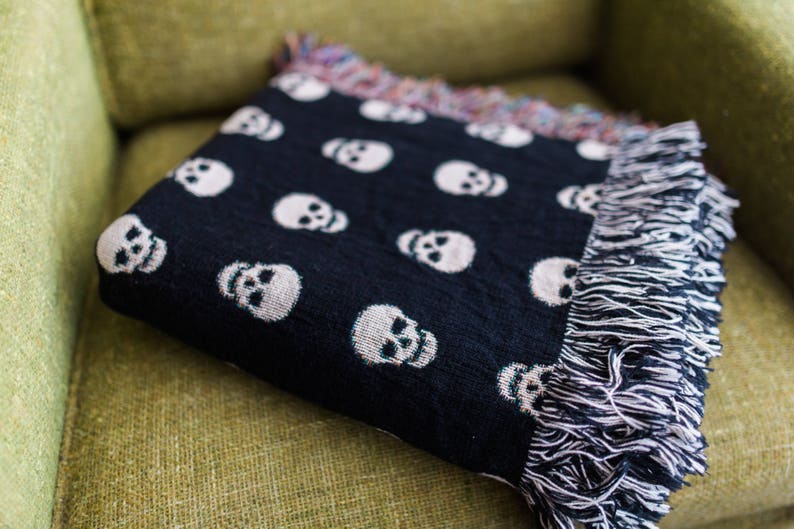 May include: A black throw blanket with a white skull pattern and fringe edges. The blanket is folded and placed on a green couch.