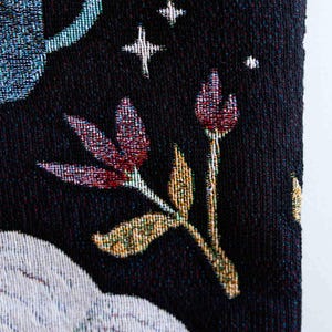 May include: A black tapestry with a white floral design and a blue crescent moon. The tapestry has a textured woven fabric.