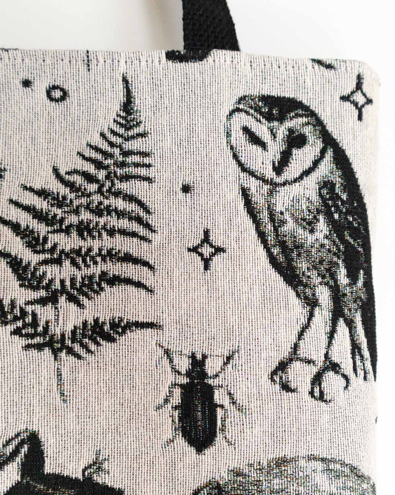 May include: A woven tote bag featuring a cream-colored fabric with a black pattern. The design includes a detailed barn owl, a coniferous tree, a small beetle, and various star shapes scattered across the surface. It has black fabric handles.