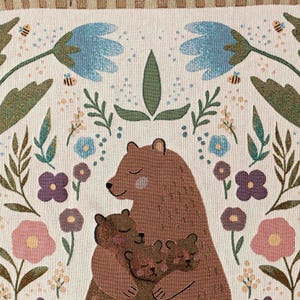 May include: A woven tapestry depicting a brown bear mother cuddling her two cubs. The background is cream with a floral design in shades of pink, purple, blue, and green.