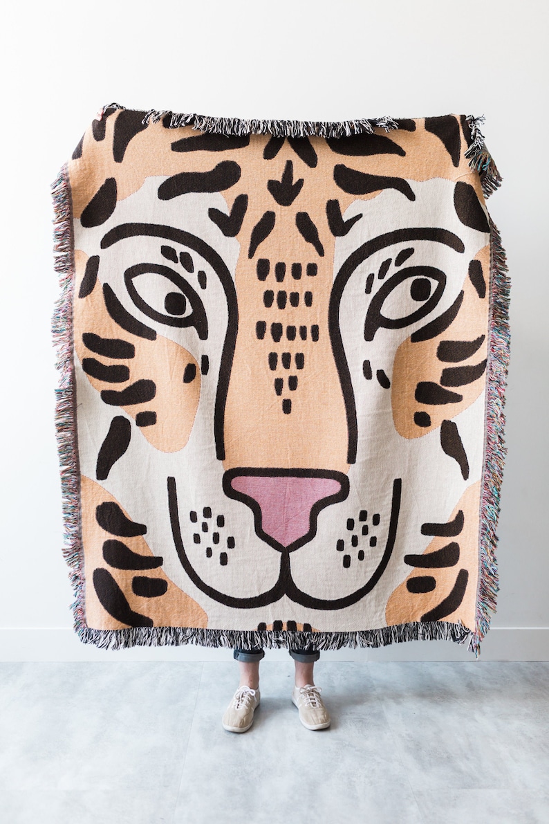 May include: A woven throw blanket featuring a large tiger face design in orange, black, white, and pink. The blanket has a fringed edge.