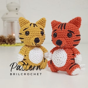 May include: Two crocheted tiger plushies, one yellow and one orange, with black stripes and white faces. The plushies are sitting on a white surface. The text "Pattern BRILCROCHET" is visible below the plushies.