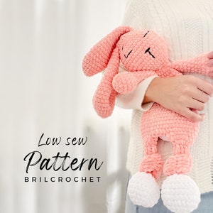 May include: A pink crocheted bunny with white feet, held by a person. The bunny has a sleepy expression and is made with a soft, fluffy yarn. The text "Low sew Pattern BRILCROCHET" is visible below the bunny.