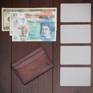 May include: A brown leather wallet with a card slot and a logo on the front. The wallet is lying on a wooden surface next to a stack of British pound notes and four blank white cards.