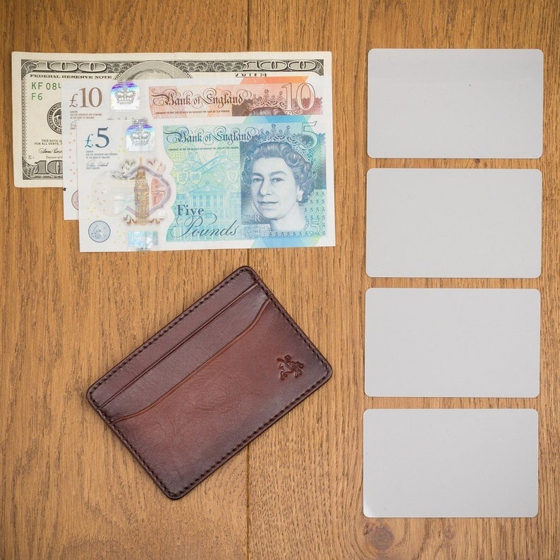 May include: A brown leather card holder with a subtle embossed design sits on a wooden surface next to a stack of British pound notes and four blank white cards.