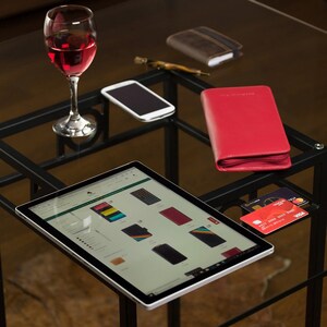 May include: A glass table displays a tablet with a website open, a red wallet, a smartphone, a pen, a brown wallet, a glass of red wine, and credit cards. The tablet shows various phone case designs.
