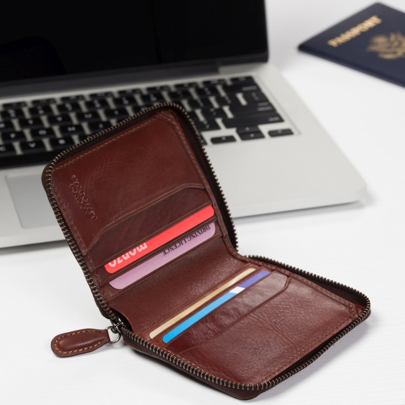 May include: A brown leather zippered wallet with a zipper pull. The wallet is open and has several cards inside. The wallet is sitting on a white surface next to a laptop.